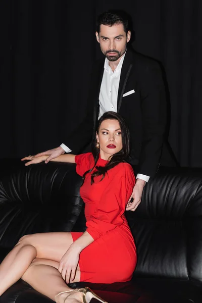 Man in suit touching sensual girlfriend in dress on leather couch on black — Stock Photo