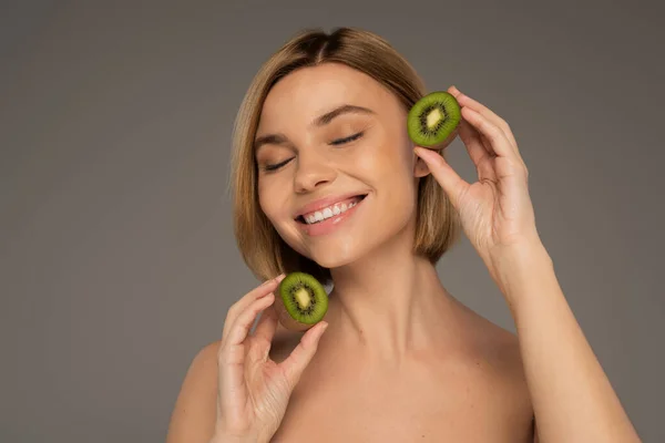 Happy young woman with closed eyes holding kiwi halves isolated on grey — Fotografia de Stock