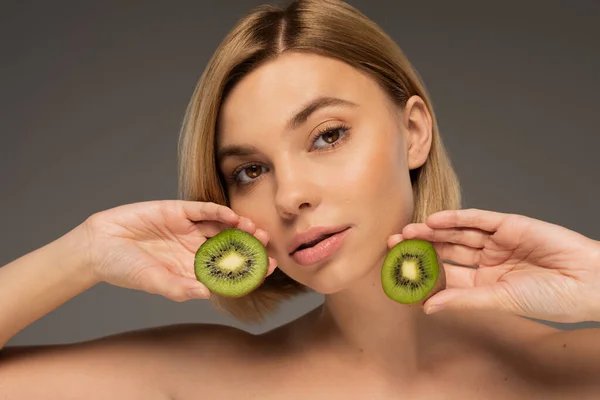Young woman with naked shoulders holding kiwi fruit and looking at camera isolated on grey - foto de stock
