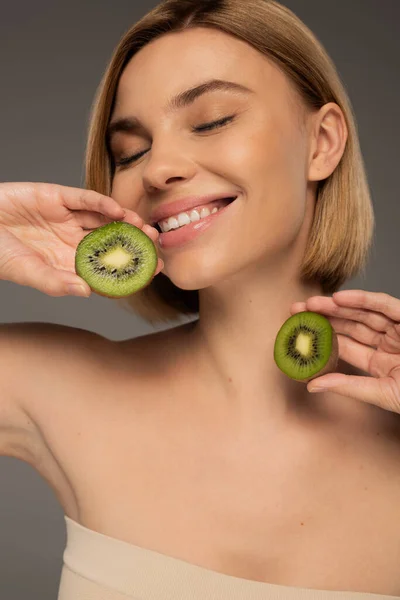 Cheerful young woman with closed eyes holding kiwi fruit halves isolated on grey — Fotografia de Stock