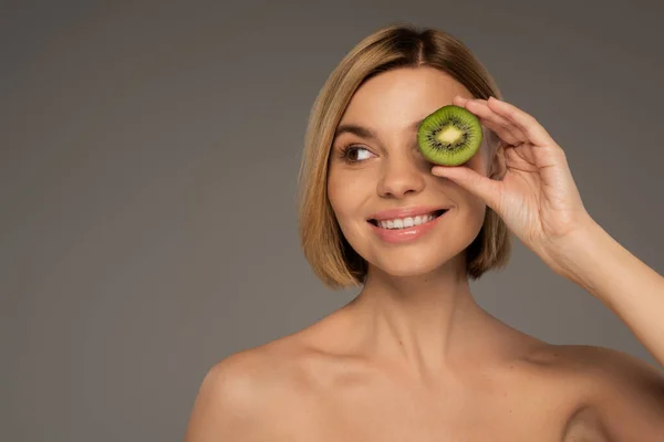 Happy young woman with bare shoulders covering eye with kiwi fruit isolated on grey — Stock Photo