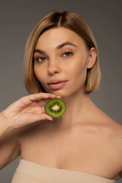 Blonde young woman with bare shoulders holding kiwi fruit and looking at camera isolated on grey — Stock Photo