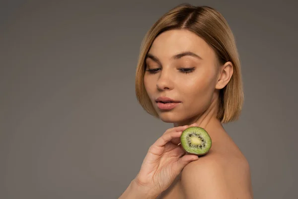 Young blonde woman with bare shoulders holding kiwi fruit isolated on grey — Stock Photo