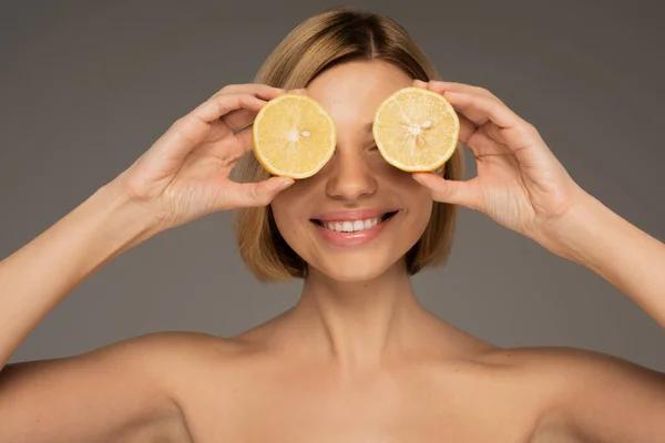 Happy young woman with bare shoulders covering eyes with lemon halves isolated on grey — Stockfoto