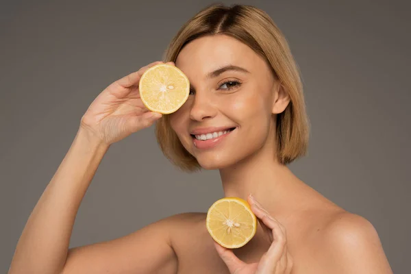 Positive woman with bare shoulders holding lemon halves isolated on grey — Stockfoto