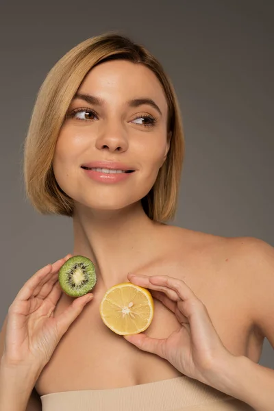 Positive woman with bare shoulders holding kiwi fruit and lemon isolated on grey — Fotografia de Stock