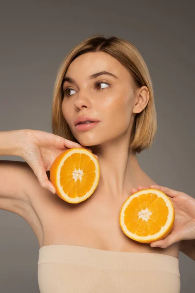 Pretty young woman with bare shoulders holding orange halves isolated on grey — Photo de stock