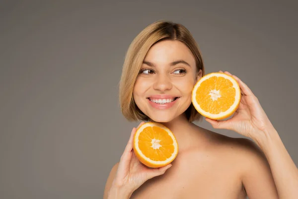 Cheerful woman with bare shoulders holding orange halves isolated on grey — Foto stock