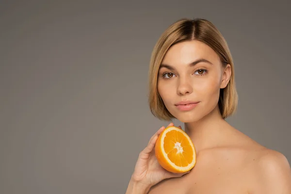 Blonde woman with brown eyes and naked shoulders holding orange isolated on grey — Photo de stock