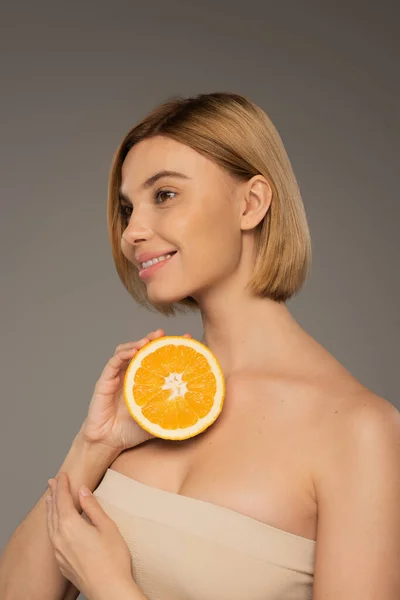 Веселая женщина с обнаженными плечами, держащая оранжевый и глядя в сторону изолированы на сером — стоковое фото