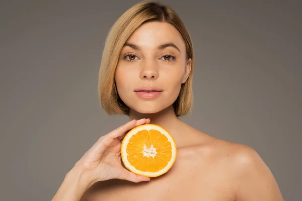 Молодая блондинка с голыми плечами, держащая апельсин и смотрящая на камеру, изолированную на сером — стоковое фото