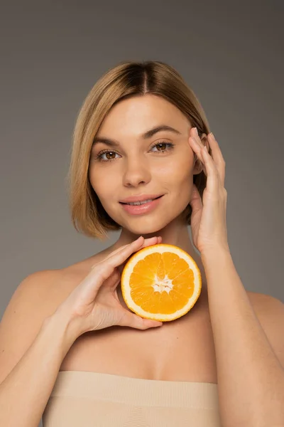 Smiling woman with bare shoulders holding juicy orange and looking at camera isolated on grey - foto de stock