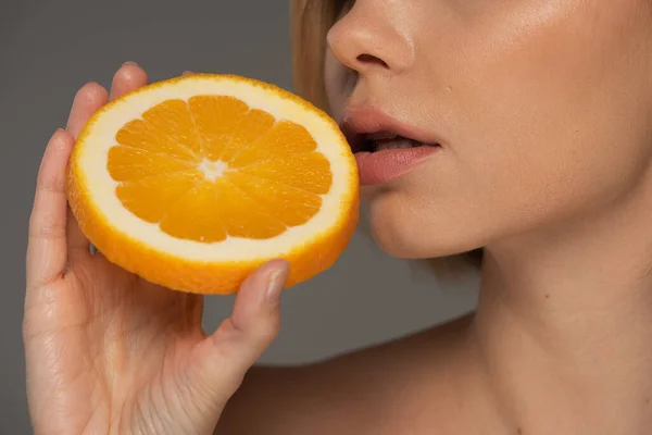 Обрезанный вид молодой женщины, держащей сочный оранжевый половину около мягких губ изолированы на серый — стоковое фото