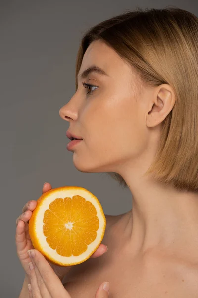 Profile of young blonde woman holding juicy orange half isolated on grey — Stockfoto