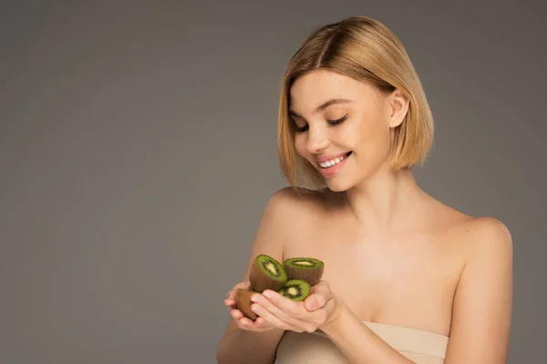 Young woman with naked shoulders looking at pile of kiwi fruit isolated on grey — Photo de stock