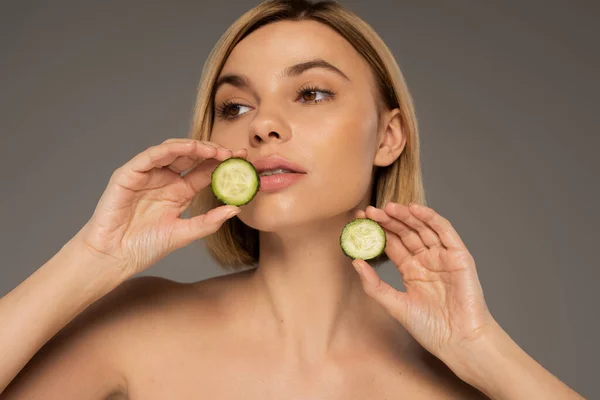 Young woman with naked shoulders holding sliced fresh cucumbers isolated on grey - foto de stock