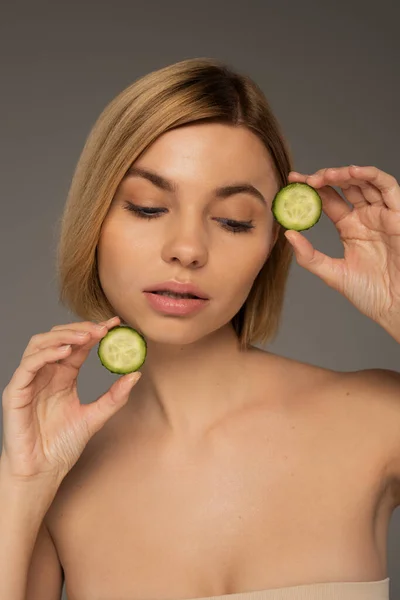 Blonde woman with naked shoulders holding sliced fresh cucumbers isolated on grey - foto de stock