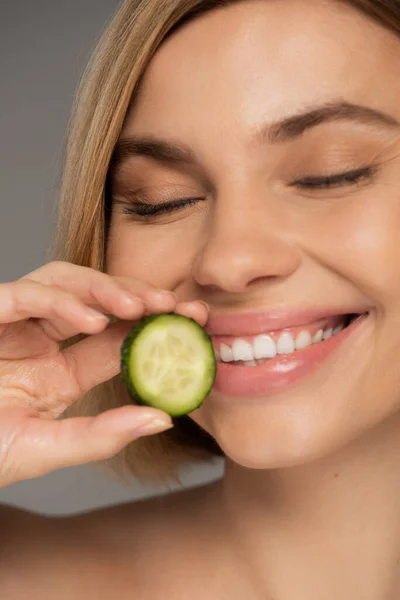 Close up of cheerful woman with closed eyes holding sliced cucumber isolated on grey - foto de stock
