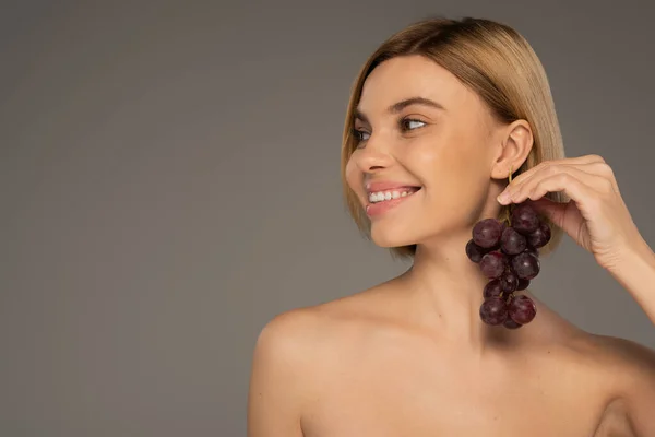 Happy young woman with bare shoulders holding ripe grapes near ear isolated on grey — Fotografia de Stock