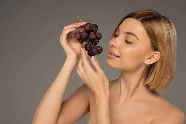 Joyful young woman with bare shoulders holding ripe grapes isolated on grey — Fotografia de Stock