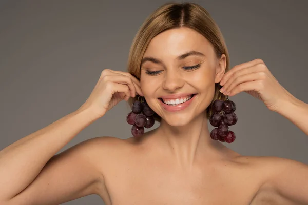 Smiling young woman with bare shoulders holding sweet grapes near ears isolated on grey — Fotografia de Stock