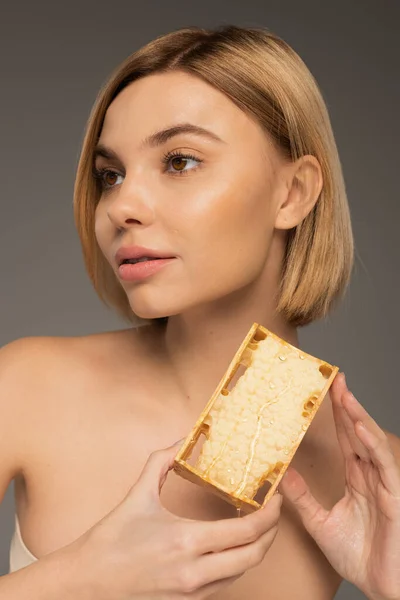 Blonde young woman with bare shoulders holding honeycomb while looking away isolated on grey — Fotografia de Stock