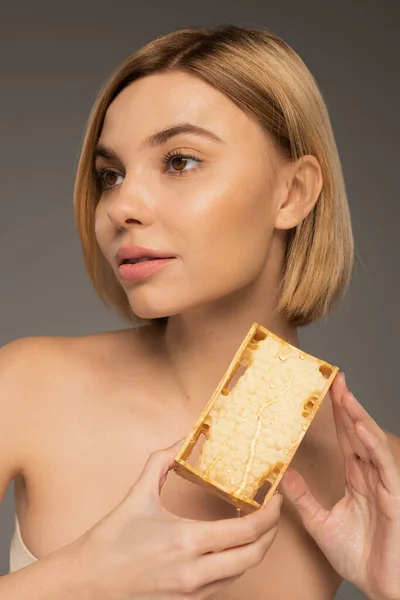 Blonde young woman with bare shoulders holding honeycomb while looking away isolated on grey — Stock Photo