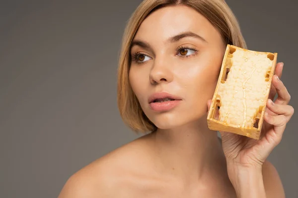 Blonde woman with bare shoulders holding natural honey in honeycomb isolated on grey - foto de stock