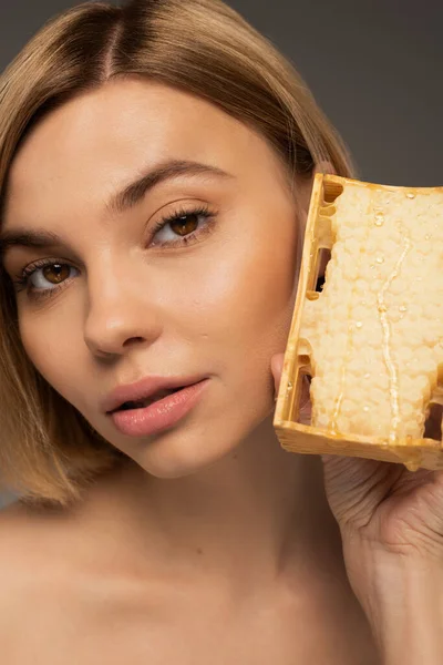 Blonde young woman with bare shoulders holding honeycomb while looking at camera isolated on grey - foto de stock