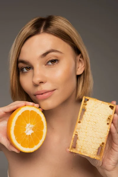 Blonde young woman holding honeycomb and orange while looking at camera isolated on grey — Fotografia de Stock