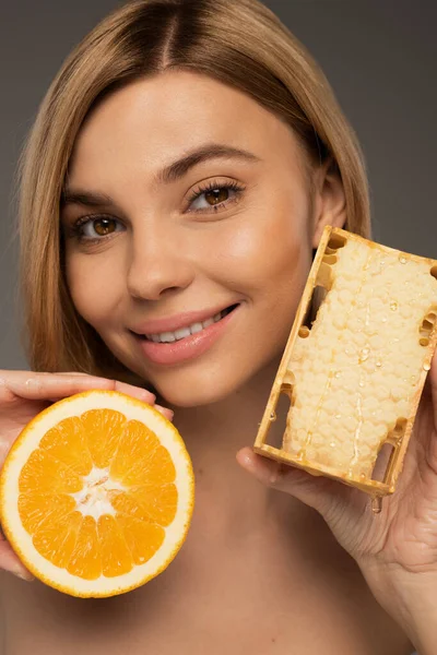 Cheerful young woman holding honeycomb and orange while looking at camera isolated on grey — Stockfoto