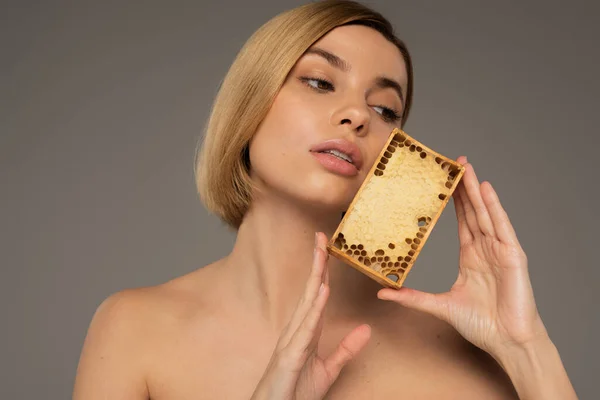 Blonde young woman holding wooden frame with sweet honey while looking away isolated on grey — Stockfoto