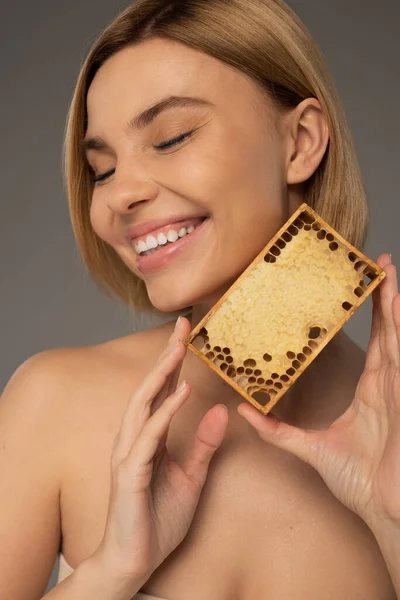 Щаслива жінка з закритими очима тримає дерев'яну раму з солодким медом ізольовано на сірому — стокове фото