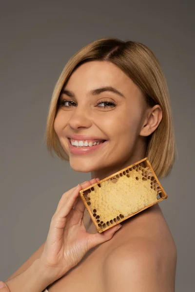 Позитивна молода жінка тримає дерев'яну раму з солодким медом ізольовано на сірому — стокове фото