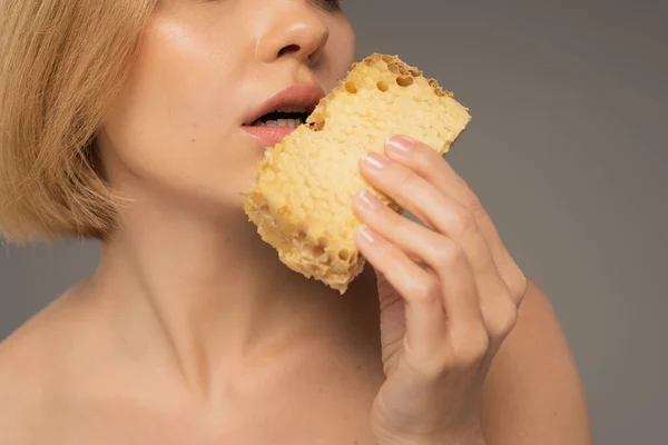 Обрізаний вид молодої жінки, що тримає шматок солодкого стільника біля рота ізольовано на сірому — стокове фото