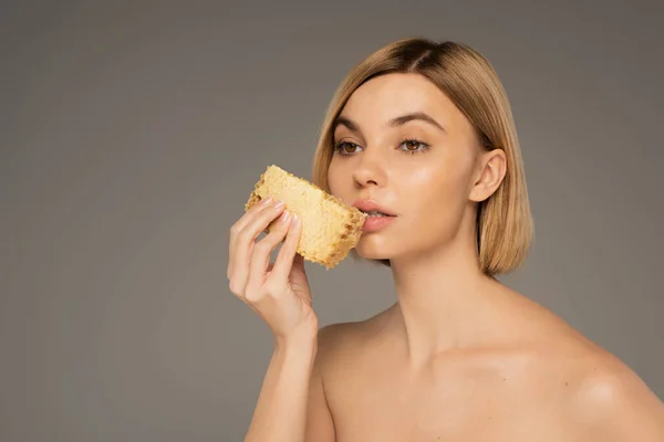 Young woman with bare shoulders holding piece of sweet honeycomb near mouth isolated on grey — Fotografia de Stock