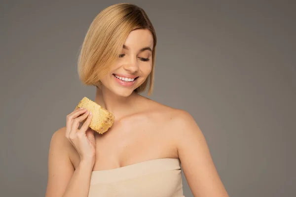 Cheerful young woman with bare shoulders holding piece of sweet honeycomb isolated on grey — Stock Photo