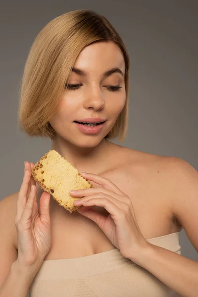 Blonde woman with bare shoulders holding piece of sweet honeycomb isolated on grey — Stock Photo