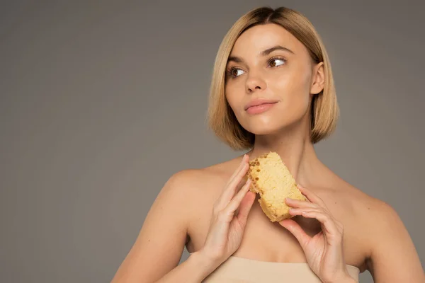 Blonde woman with bare shoulders holding piece of sweet honeycomb while looking away isolated on grey — Foto stock