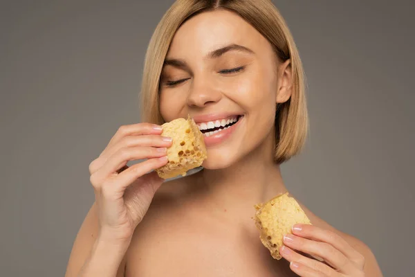 Joyful woman with bare shoulders holding pieces of sweet honeycomb isolated on grey — Foto stock