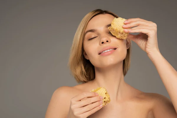 Pleased young woman with bare shoulders holding sweet honeycomb near eye isolated on grey - foto de stock
