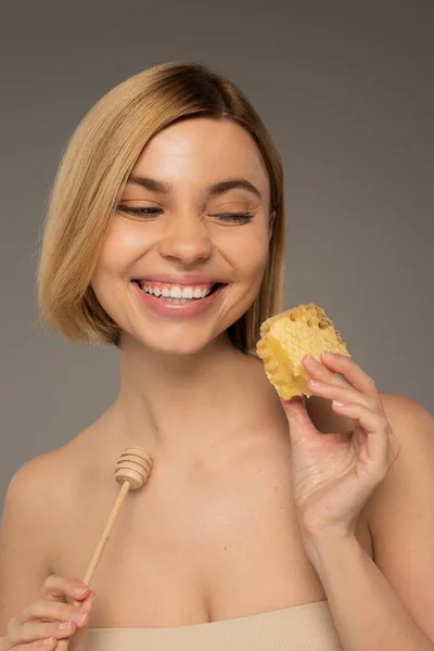 Pleased young woman with naked shoulders holding sweet honeycomb and wooden dipper isolated on grey - foto de stock