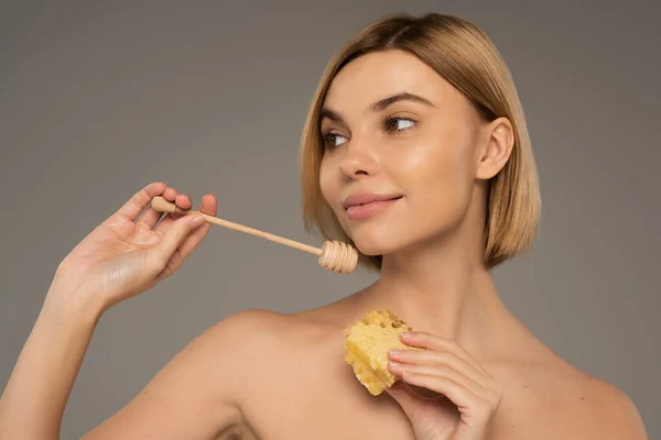 Pleased woman with bare shoulders holding sweet honeycomb and wooden dipper isolated on grey — Foto stock