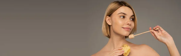 Pleased young woman with naked shoulders holding sweet honeycomb and wooden dipper isolated on grey, banner — Stock Photo