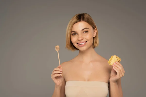 Happy woman with naked shoulders holding sweet honeycomb and wooden dipper isolated on grey — Foto stock