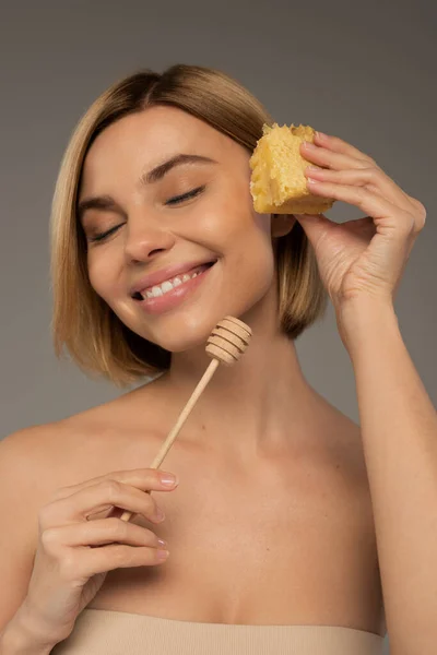 Smiling woman with closed eyes holding sweet honeycomb and wooden dipper isolated on grey — Stock Photo