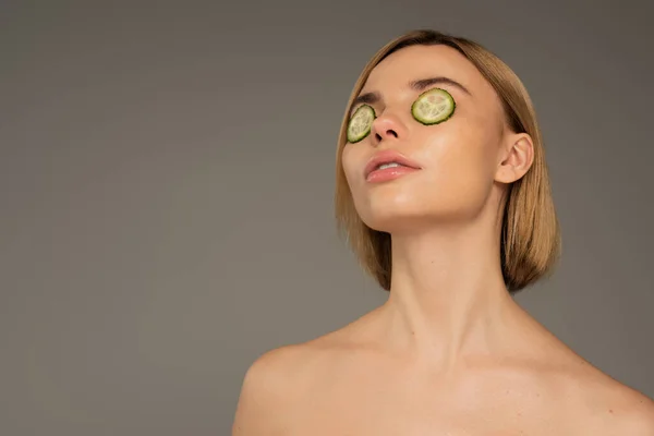 Blonde young woman with naked shoulders and fresh cucumbers on eyes isolated on grey — Photo de stock