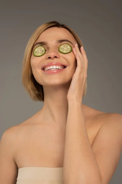 Joyful young woman with naked shoulders and fresh cucumbers on eyes isolated on grey - foto de stock