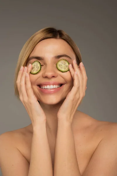 Cheerful young woman with naked shoulders and fresh cucumbers on eyes isolated on grey - foto de stock