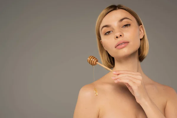 Blonde woman holding dipper and pouring sweet honey on naked shoulder isolated on grey — Foto stock